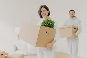 Happy young couple enter own modern house, buy real estate, carry cardboard boxes with indoor plant and other personal stuff, buy new house for living, have moving day. People and property concept photo