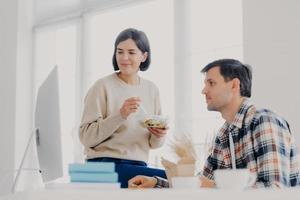 Female and male colleagues collaborate together, eat salad, have lunch time, concentrated into computer, pose in coworking space, search necessary information and prepare project for meeting photo