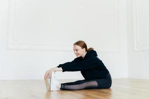 Fit redhead woman stretches muscles on legs, has sporty body, focused into distance, looks happily, wears black clothes, poses on floor in spacious hall. People, flexibility and sport concept photo