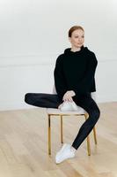 Vertical shot of thoughtful red haired woman with make up, takes rest on chair, wears black sweatshirt, leggings, white sneakers, focused away, thinks about something. People and sport concept photo