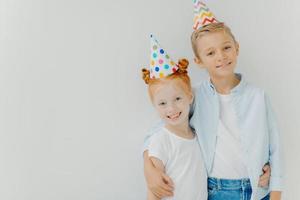 Happy small ginger freckled girl and little boy embrace and smile gladfully, wear party hats, enjoy nice time together, isolated over white background, copy space. Children and festive event concept photo