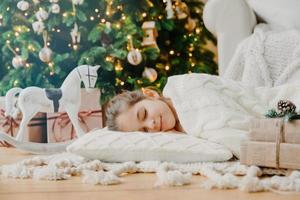 Charming girl sleeps on soft white pillow on floor against decorated New Year tree, has pleasant dreams, surrounded with toy horse and gift boxes. Children, rest and winter holidays concept. photo