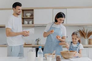 Mother and father give eggs to daughter who prepares dough, busy cooking together during weekend, have happy moods, prepare food. Three family members at home. Parenthood and togetherness concept photo