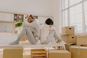 Photo of stressful couple get divorse, leave house, start living seperately, sit backs to each other, pose in kitchen, cardboard boxes with belongings around, focused down with sad expressions