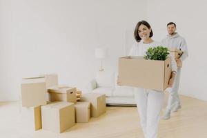 Happy woman and her husband carry boxes with personal belongings, being busy during relocation in other place for living, enter new home, move together in big house, unpack household things. photo