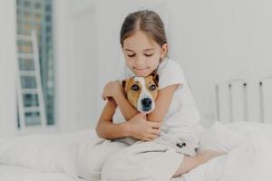 Indoor shot of happy small child embraces favourite pet, dressed in pyjamas, expresses love to her dog, plays with animal after awakening, poses on soft bed. Children and domestic animals concept photo