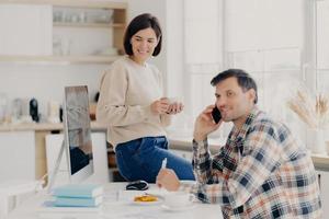 Horizontal shot of cheerful man has telephone conversation, discusses financial contract agreement, dressed in checkered shirt, writes down information, happy woman holds mug of drink, leans at table photo