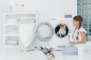 Positive small girl has fun with jack russel terrier, poses on floor near washing machine in laundry room, holds liquid powder, busy with housework, has glad expression. Cozy interior in bathroom photo