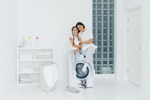 Happy mother and daughter have fun, embrace and express love to each other, dressed in casual wear, sit on washer, prepare for washing, being in white spacious laundry room, basin with dirty clothes photo