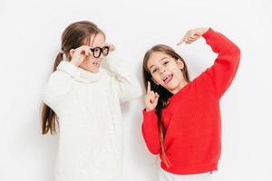 Cheerful two sisters have fun together, have funny expressions. Adorable child in eyewear and white warm sweater looks at her best female friend who makes grimace, isolated over studio background photo