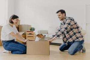 Funny young woman and man have fun during relocation, pose on floor with dog in cardboard box, just moved into apartment, dressed in domestic clothes, sofa and pile of containers in background photo