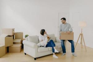 Positive woman and man pose in empty spacious room during relocation day, husband carries cardboard boxes with belongings, wife has telephone conversation, sits on comfortable sofa with dog. photo