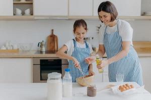 Brunette mother adds oil in dough, little daughter helps to make pastry, whisks ingredients, pose together against kitchen interior, prepare bakery together. Small helper with mommy. Homemade food photo