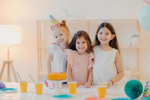 Indoor shot of happy three girls embrace and have fun, smile gladfully, stand near festive table with cake, cups, have happy childhood, being on party together. Childhood and festivity concept photo