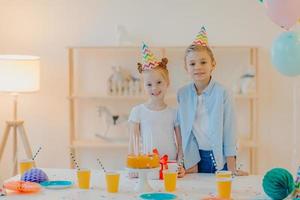 Glad boy and his small ginger sister dressed in festive clothes, party hats, celebrate birthday together, surrounded with cake, present and cups of drink, have good mood during special occasion photo