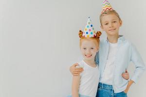 una foto aislada de un hermano y una hermana felices se abrazan, tienen expresiones positivas, usan sombreros de fiesta, van a celebrar el cumpleaños, se paran contra el fondo blanco, copian el espacio a un lado. niños alegres