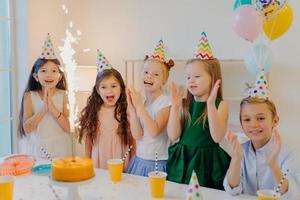 Cheeful friends of five pose near festive table with big cake, cup of drinks, wear party hats, look joyfully at sparkle, laugh happily, pose indoor, celebrate birthday photo