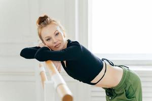 Restful fatigue graceful female dancer practices in dance studio, looks at camera with tired expression, leans at horizontal bars for performing exercises, wears black top and sweats, takes break photo