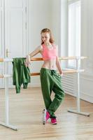 Beautiful fit ginger female dancer has rest near barre in ballet dancing studio, focused down with thoughtful expression, wears top, green trousers and sneakers, prepares for performance on stage