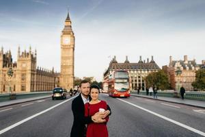 una encantadora pareja casada se abraza en la carretera con el fondo del big ben y el autobús londinense, pasan vacaciones juntos en gran bretaña, reciben emociones agradables y buenas impresiones foto