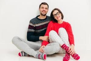 Horizontal shot of happy lovers sit together against white studio background, wear casual clothes, enjoy leisure time at home. Adorable young female and male partners have pleasant smiles on face photo