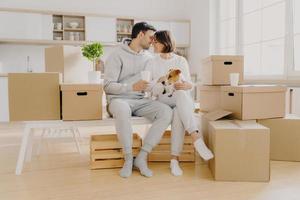 Moving to new home, family time concept. Couple in love going to kiss each other, sit at white bench against kitchen interior, play with dog, surrounded with many packages, enjoy first day in new home photo