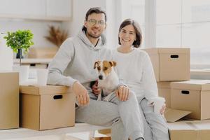 Positive woman and man pose near stack of cardboard boxes, pose for making portrait with dog, relocate in new living place, wear white casual clothes, pose in spacious kitchen with big windows photo