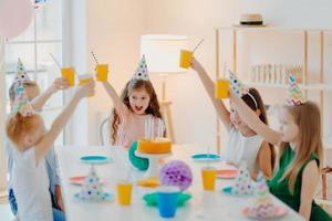 Group of cheerful preschool kids celebrate birthday together, have fun, cheer with cups of beverage, wears festive hats, eat delicious cake, sit at table in spacious room. Children and party concept photo