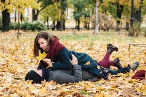 People, leisure and season concept. Happy female and male enjoy spending time together, lie on ground covered with yellow leaves. Cheerful girlfriend and boyfriend look happily at each other photo