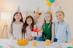 Glad birthday girl stands with presenet, happy friends come to congratulate her, wear party cone hats, stand near festive table with cake, smile joyfully, celebrate festive event photo