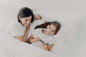 Caring mom looks attentively at her little daughter who wakes up early, listens music in earphones, searches new song in playlist of modern smartphone, lie under soft white blanket. Top view photo