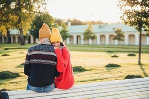 Beautiful female in loose red sweater embraces her boyfriend, missed him too much as didnt see long ago, sit on bench in park, have pleasant conversation, admire wonderful landscape or nature photo