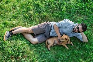 Attractive European man in casual clothing and sunglasses is lying on grass and hugging his cute little dog. photo