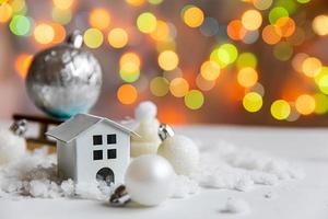 Abstract Advent Christmas Background. Toy model house and winter decorations ornaments toys and balls on background with snow and defocused garland lights. Christmas with family at home concept. photo