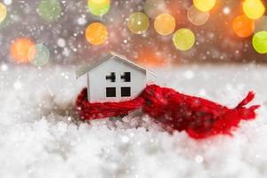 Abstract Advent Christmas Background. Toy model house with knitted red scarf on snow with defocused garland lights background. Christmas with family at home concept. Christmas winter composition. photo