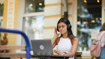 Woman sits at outdoor cafe using laptop wearing headphones