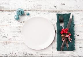 Christmas table setting with white dishware, silverware and red and green decorations on white wooden background. Top view. photo