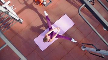 une jeune femme athlétique dans un survêtement de sport violet fait des exercices d'étirement sur un terrain d'entraînement dans un parc de la ville. remise en forme à l'extérieur. ralenti. vue de dessus video