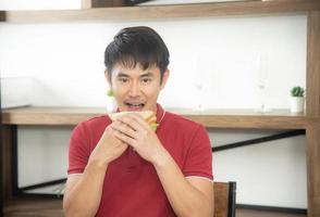 el hombre sonriente de negocios con una camiseta roja informal desayunando y comiendo un sándwich, un joven cocinando comida y bebida en la cocina estilo loft foto