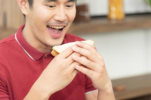 Asian smiling young man with casual  red t-shirt enyoy having breakfast, eating sandwich, Young man cooking food and drink in the loft style kitchen room photo