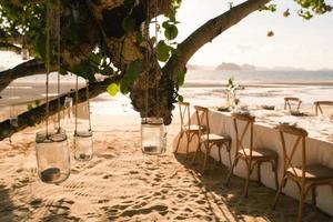 Close up the jar candle hanging on the tree with long table wedding dinner setup on the beach at Thailand in the evening. Wedding party concept. Decoration outdoor restaurant at the beach. photo