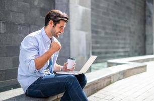 joven caucásico sentado y trabajando en un proyecto independiente usando una computadora portátil en la oficina exterior. sintiéndose feliz mientras su trabajo acaba de tener éxito. foto