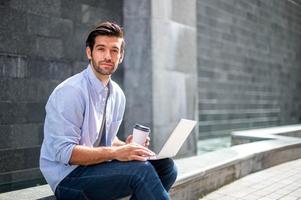 Young caucasian man looking camera while working on freelance project using portable computer at outer office. photo