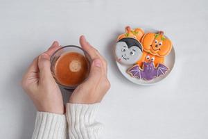 mano de mujer sosteniendo la taza de café durante la comida divertidas galletas de halloween. feliz día de halloween, truco o amenaza, hola octubre, otoño otoño, tradicional, concepto de fiesta y vacaciones foto