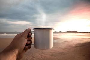 Cafe mañanero. bebiendo café caliente en la playa durante el amanecer. mano sosteniendo una taza para disfrutar de la bebida favorita y la naturaleza. comenzar un nuevo día. estilo de vida relajante y armónico. vista pov foto