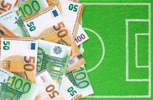 Euro banknotes 50 and 100 placed on small football field made of green felt, top view. Sports betting, soccer bets, football match results, gambling, money. Background with copy space. photo