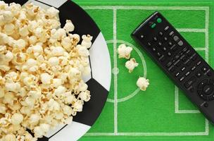 Football soccer party table. Popcorn on big plate painted like soccer ball and dish mat like football field made of green felt, top view. TV remote for watching foorball match broadcast. photo