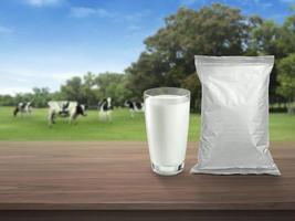 Fresh milk in glass, and foil food packaging on dark wooden tabletop, blurred background with cows on the meadow. healthy eating photo
