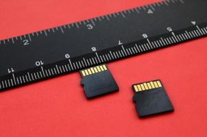 Two small micro SD memory cards lie on a red background next to a black ruler. A small and compact data and information store photo