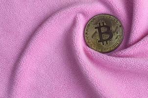 The golden bitcoin lies on a blanket made of soft and fluffy light pink fleece fabric with a large number of relief folds. The shape of the folds resembles a fan from a video card cooler photo